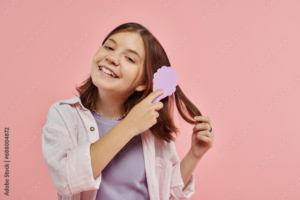 cheerful preteen girl in stylish clothes brushing hair and looking at ...