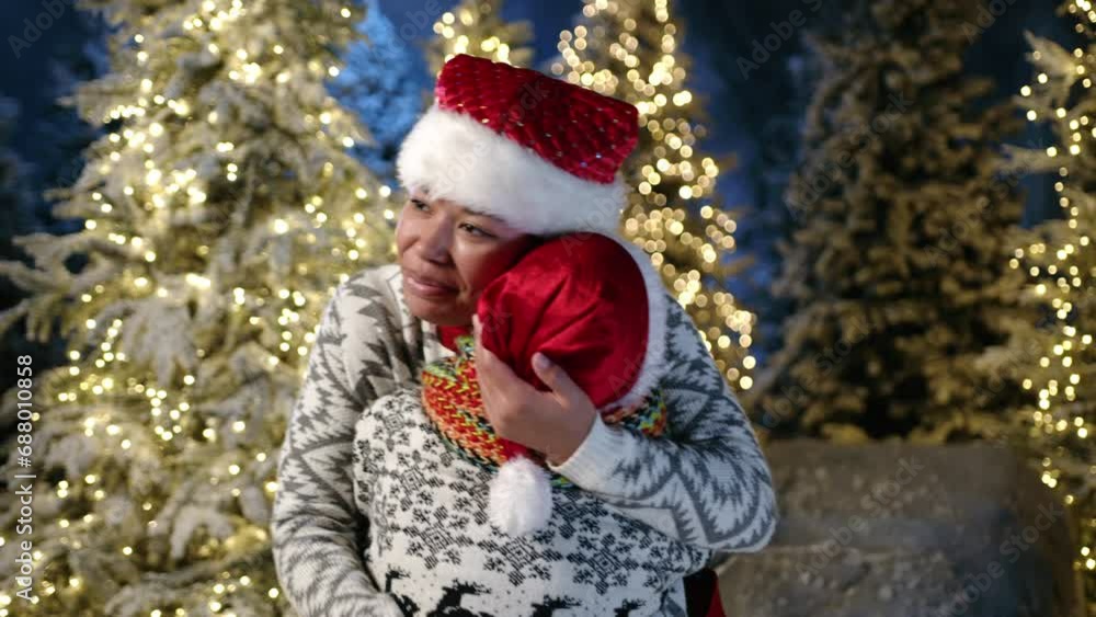 Caring mum in santa hat reaching out to kid in Christmas outfit, hugging, looking with love. Many conifers dusted with snow and decorated with garlands. High quality 4k footage Caring mum in santa hat reaching out to kid in Christmas outfit, hugging, looking with love. Many conifers dusted with snow and decorated with garlands. High quality 4k footage