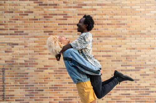 Cheerful transgender woman lifting boyfriend in front of brick wall photo