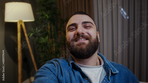 Selfie of smiling young adult man holding a smartphone in hand looking at camera, taking fhoto photo