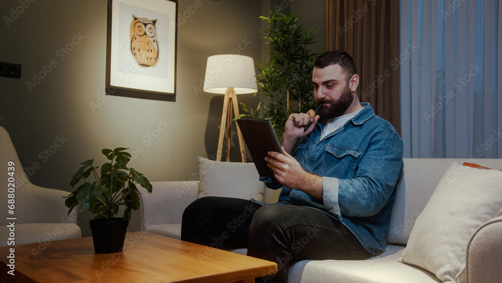 Bearded young college student working with tablet device while sitting on sofa at home at night