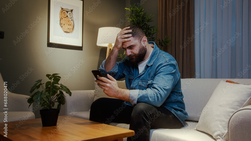 Young adult man sitting on couch at home while holding a smartphone having anxiety and stress. Upset young man reacting to loss, bad news