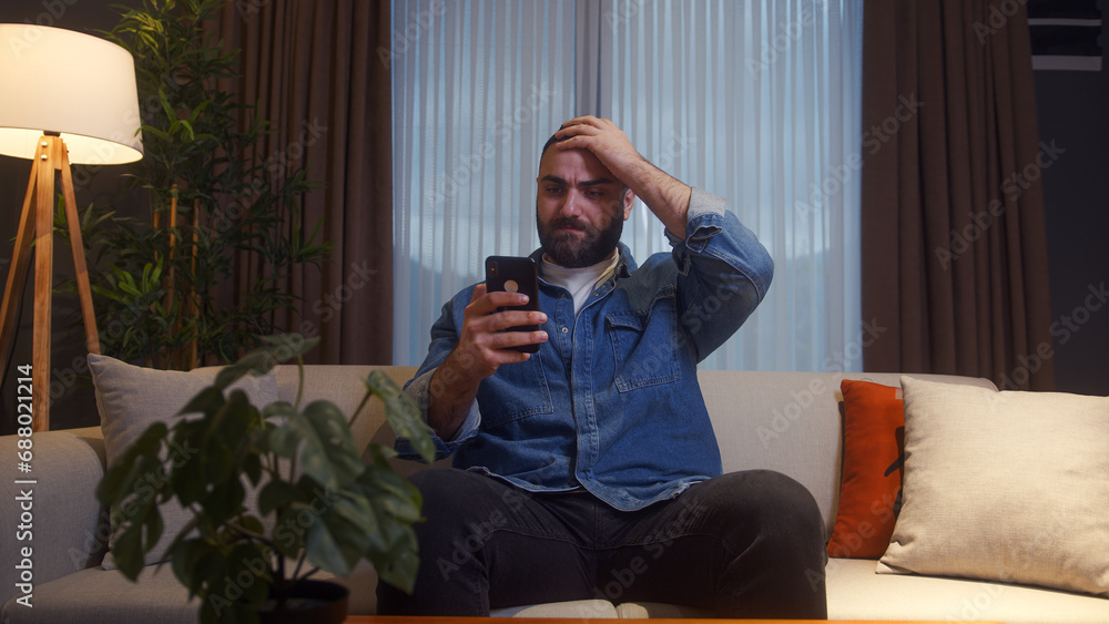 Bearded young adult man sitting on couch at home while holding a smartphone having anxiety and stress. Upset young man reacting to loss, bad news Bearded young adult man sitting on couch at home while holding a smartphone having anxiety and stress. Upset young man reacting to loss, bad news