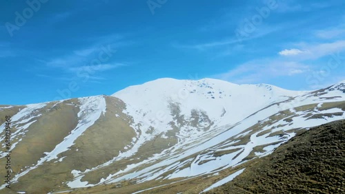Aerial view of high mountains covered with snow with thawing on a sunny day against a blue sky. Deserted gray mountains in winter without grass with snowy peaks. Drone footage. Direct flight. Armenia