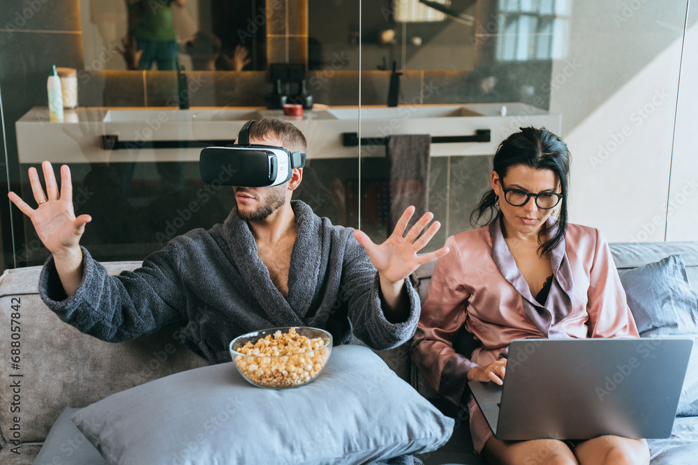 Bearded young man in bath robe sitting in bed using virtual reality headset while his wife works using laptop. Domestic leisure, gaming, emote work. Guy playing games home. Woman with computer. - obrazy, fototapety, plakaty 