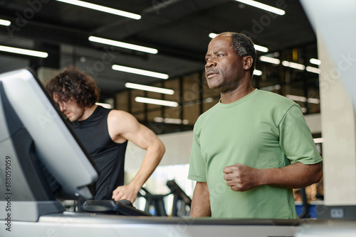 Elderly black man standing on treadmill at gym with caucasian gym goer working out on background