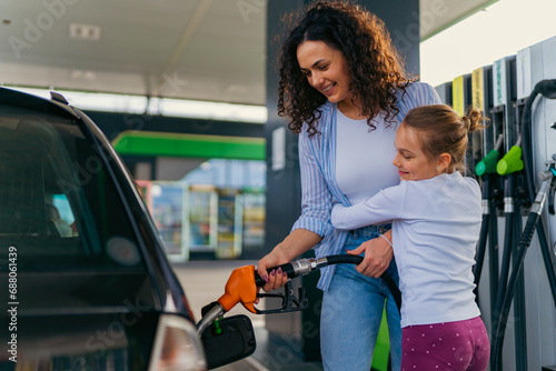 A mother pours fuel into the car while her daughter hugs her, both are happy