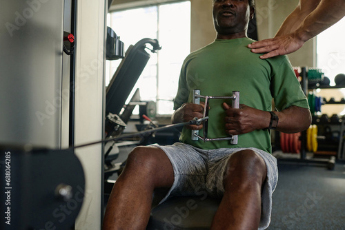 Black sportsman making low cable row at gym with unknown caucasian trainer holding his shoulders