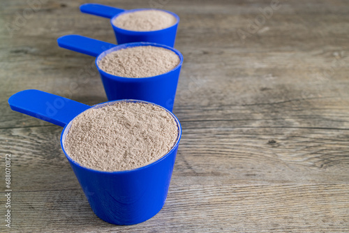 Three supplement scoops in a row filled with protein powder on top of wooden kitchen table. Dietary and athletic performance nutrition concept.