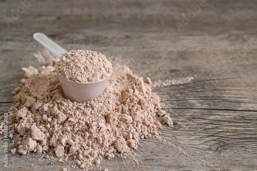 Heaping measuring scoop filled with protein shake powder sitting on a pile of protein powder spilled on wooden table surface. Fitness diet and athletic performance nutrition concept.