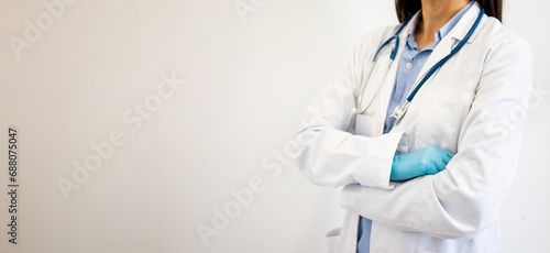 An unrecognizable woman in a white coat, latex gloves and a stethoscope poses with her arms crossed against a white studio background. Copyspace for medical advertising. Health insurers.