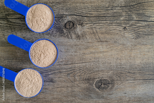 Three supplement scoops in a row filled with protein powder on top of wooden kitchen surface. Dietary supplements concept – flat lay, top view with copy space.