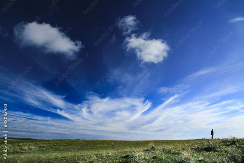Cloud swirl in a bright blue summer sky over a green meadow with one person looking tiny on the horizon Cloud swirl in a bright blue summer sky over a green meadow with one person looking tiny on the horizon