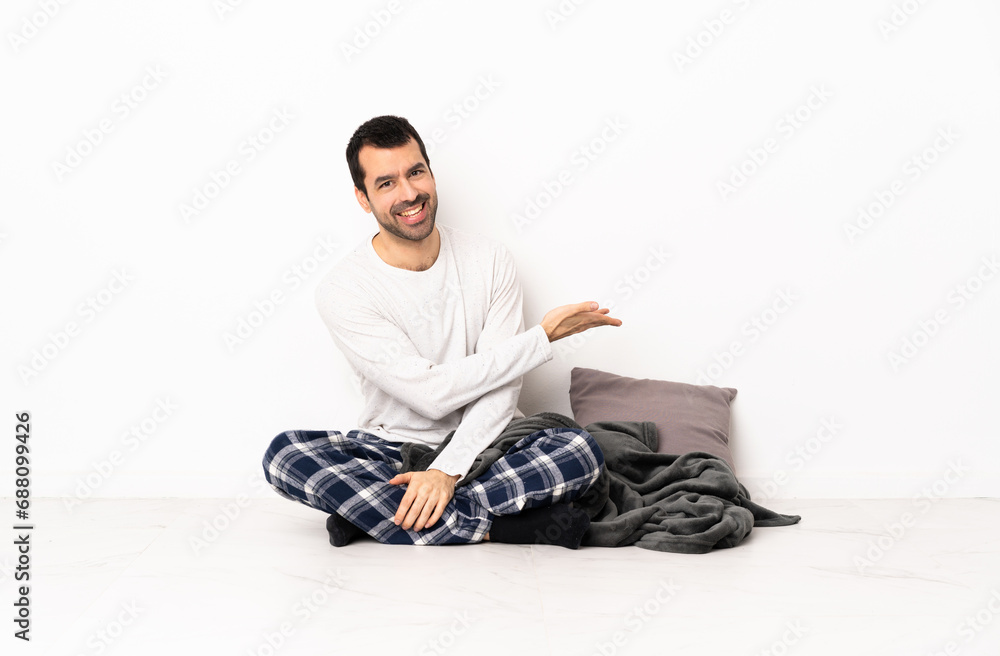 Caucasian man in pajamas sitting on the floor at indoors extending hands to the side for inviting to come Caucasian man in pajamas sitting on the floor at indoors extending hands to the side for inviting to come