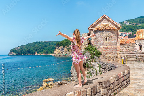 Young woman stands with her arms raised up on a parapet in the Old Town of Budva on the backdrop of a seascape - view from the back, Montenegro. Female tourist admires the beautiful summer landscape
