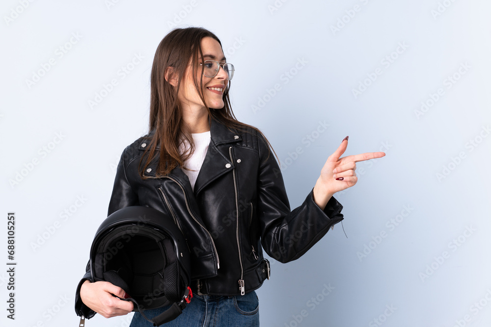 Young woman holding a motorcycle helmet over isolated blue background pointing finger to the side Young woman holding a motorcycle helmet over isolated blue background pointing finger to the side