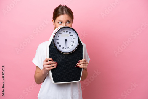 Young caucasian woman isolated on pink background with weighing machine and hiding behind it