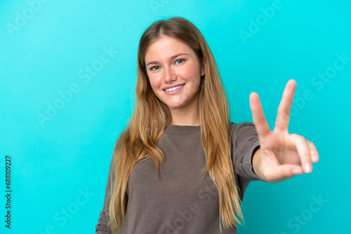 Young blonde woman isolated on blue background smiling and showing victory sign