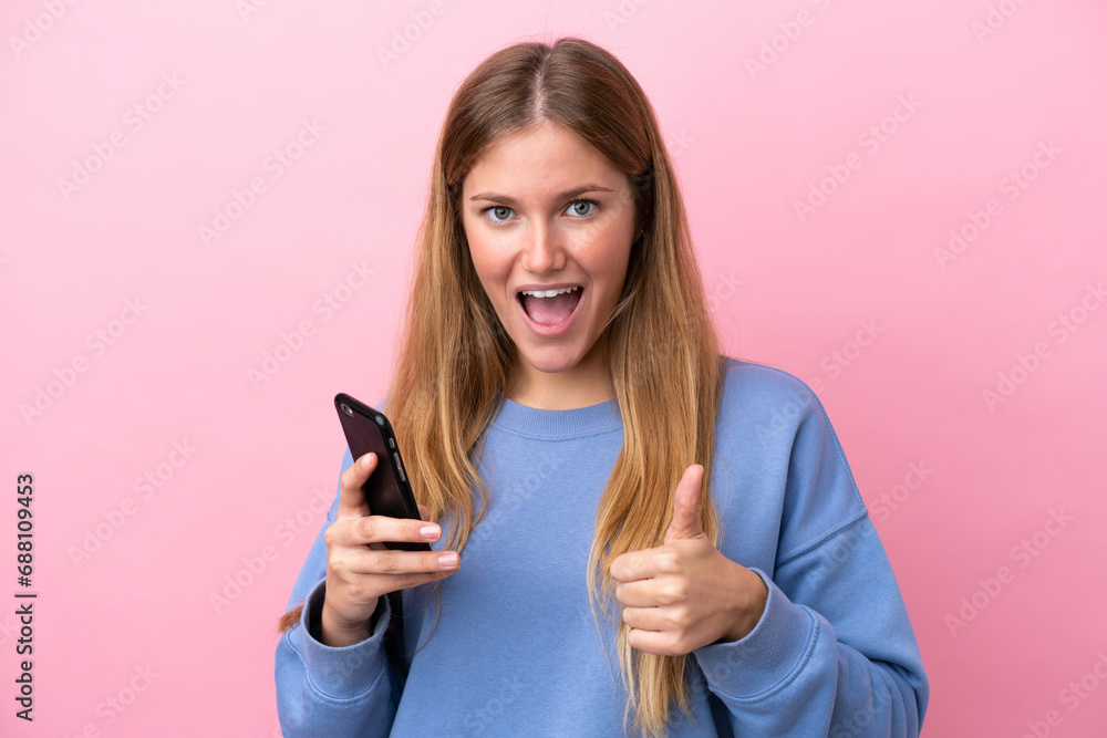 Young blonde woman isolated on pink background using mobile phone while doing thumbs up Young blonde woman isolated on pink background using mobile phone while doing thumbs up