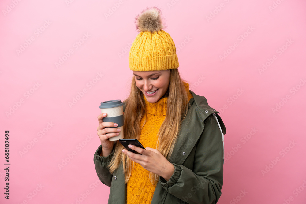 Young blonde woman wearing winter jacket isolated on pink background holding coffee to take away and a mobile - obrazy, fototapety, plakaty 