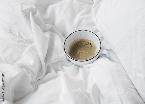 A white cup of coffee stands on a bed in white linen
