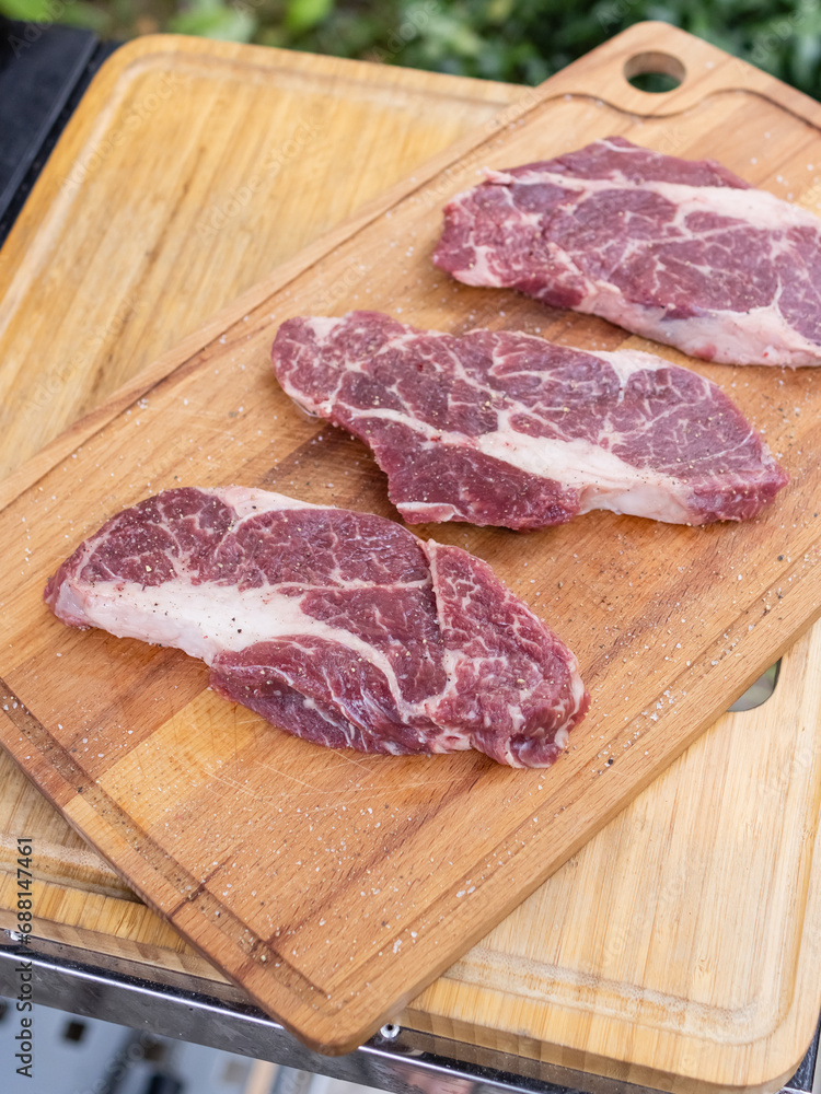 Three pieces of ribeye steak lie on a cutting board next to the grill