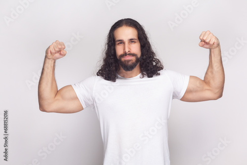 Portrait of strong powerful bodybuilder man in white t-shirt with long wavy hair and beard, standing raised his arms, showing his biceps. Indoor studio shot isolated on gray background.