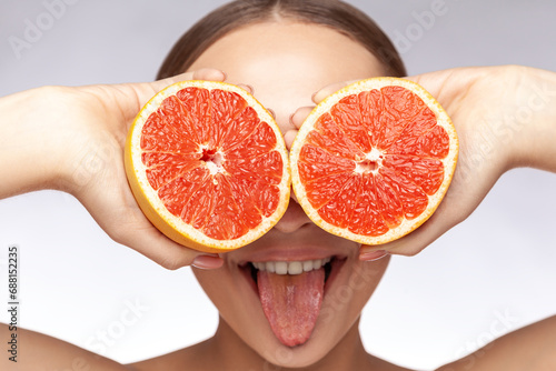 Closeup portrait of funny cheerful childish beautiful woman covering eyes with grapefruits showing tongue out. Indoor studio shot isolated over gray background.