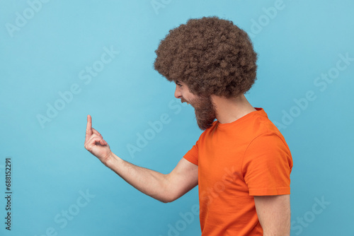 Side view of angry man with Afro hairstyle in orange T-shirt showing middle finger and asking to get off expressing negativity, disrespectful behaviour. Indoor studio shot isolated on blue background. photo