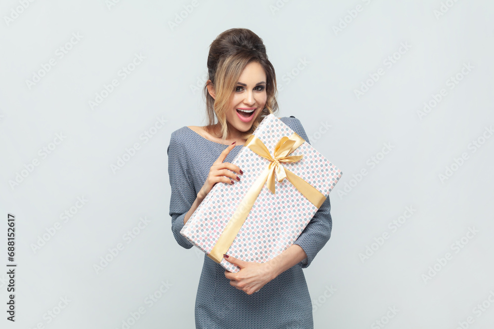 Portrait of amazed happy blonde woman with gift box, being pleasantly surprised with present for her birthday, wearing grey elegant dress. Indoor studio shot isolated on gray background.