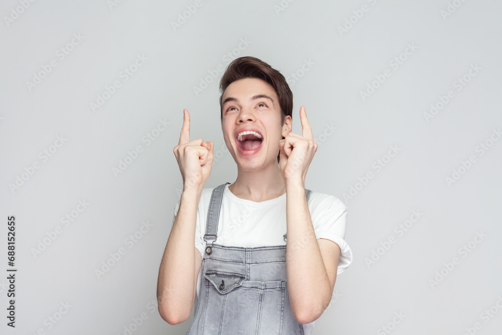 Portrait of joyful young brunette man standing looking up, pointing finger at copy space above her head, rejoicing, wearing denim overalls. Indoor studio shot isolated on gray background. Portrait of joyful young brunette man standing looking up, pointing finger at copy space above her head, rejoicing, wearing denim overalls. Indoor studio shot isolated on gray background.