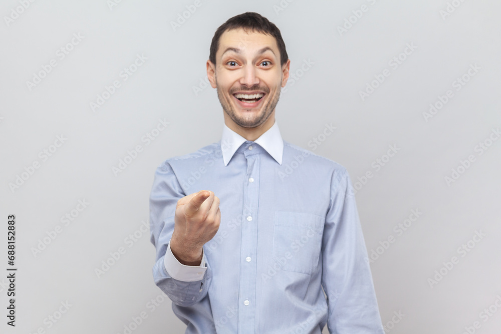 Portrait of smiling optimistic joyful attractive man standing looking at camera with toothy smile, pointing at you, wearing light blue shirt. Indoor studio shot isolated on gray background. Portrait of smiling optimistic joyful attractive man standing looking at camera with toothy smile, pointing at you, wearing light blue shirt. Indoor studio shot isolated on gray background.
