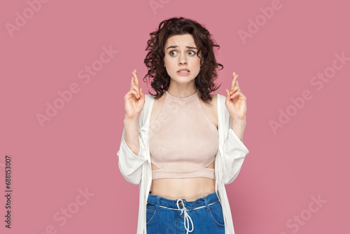 Portrait of hopeful attractive woman with curly hair wearing casual style outfit crossing her finger for good luck, waiting for miracle. Indoor studio shot isolated on pink background. photo