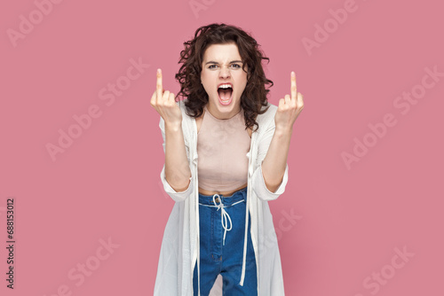 Portrait of rude impolite woman with curly hair wearing casual style outfit standing showing middle fingers, screaming with hate and anger. Indoor studio shot isolated on pink background. photo