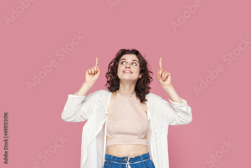 Portrait of smiling satisfied woman with curly hair wears casual style outfit pointing up with both index fingers, showing copy space for advertisement. Indoor studio shot isolated on pink background.