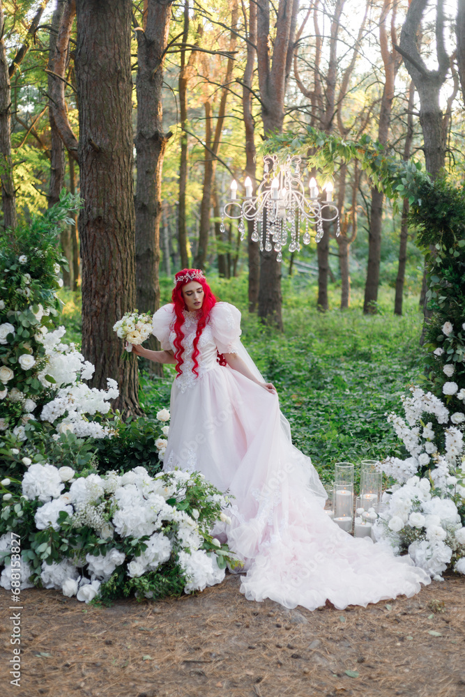 Beautiful couple, the groom in a green suit and the bride in an unusual pink wedding dress, posing at a luxurious ceremony with beautiful decor and an arch with a chandelier and candles in the forest 