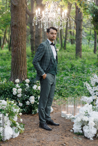 Beautiful couple, the groom in a green suit and the bride in an unusual pink wedding dress, posing at a luxurious ceremony with beautiful decor and an arch with a chandelier and candles in the forest 