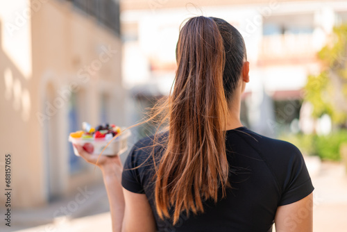 Young pretty brunette woman holding a bowl of fruit at outdoors in back position