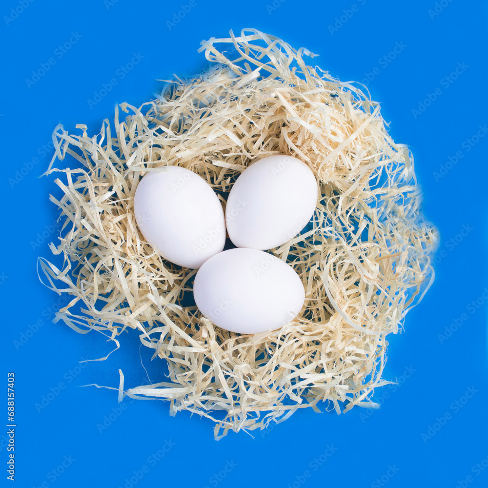 White easter eggs in a nest of straw on the blue background. Close-up. White easter eggs in a nest of straw on the blue background. Close-up.