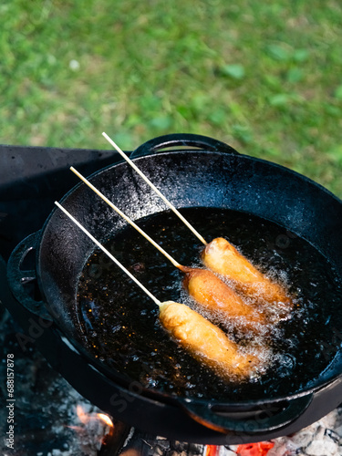 Several corn dogs on sticks are cooked in hot oil in a cast iron cauldron