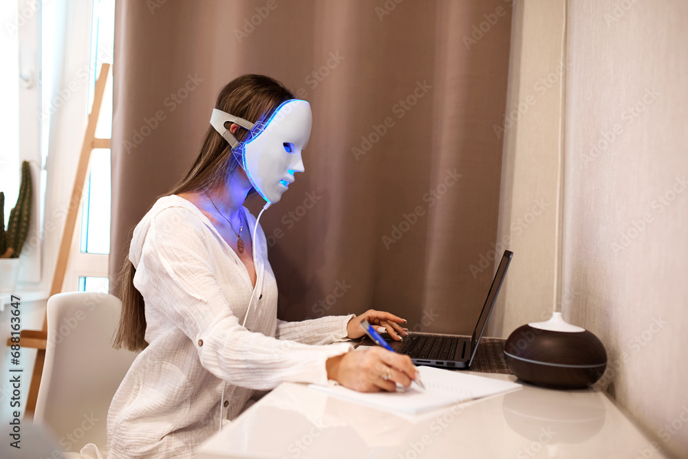 Naklejka premium A beautiful girl with an LED mask on her head works at a laptop. Home skin care concept.