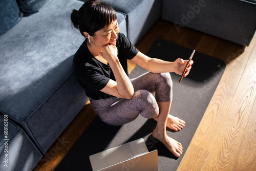 Smiling young woman sitting on yoga mat using smartphone at home photo