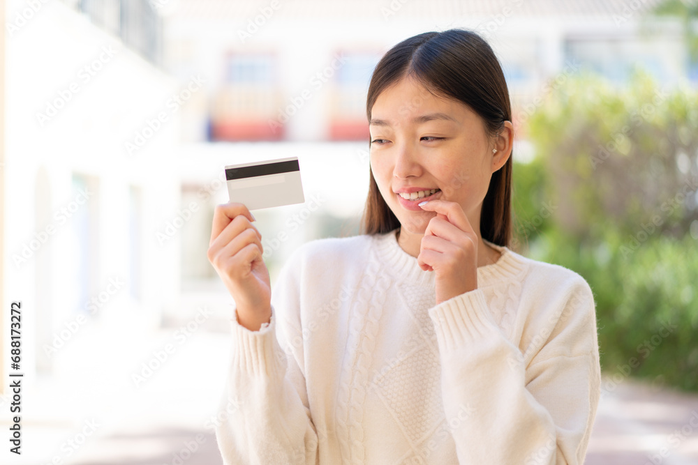 Pretty Chinese woman at outdoors holding a credit card and having doubts Pretty Chinese woman at outdoors holding a credit card and having doubts
