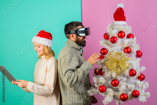 Bearded man in VR headset and pretty girl in Santa hat with laptop near Christmas tree. Modern technology and entertainment. Christmas couple playing games at home and using VR glasses and notebook.