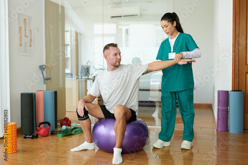 Patient of rehabilitation center sitting on fitness ball when doing arm exercise under control of nurse