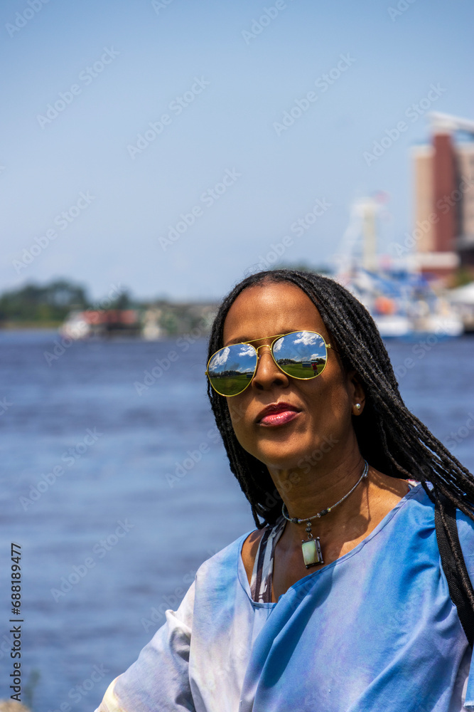 An African American woman with long sisterlocks wearing a colorful dress and sunglasses along Cape Fear river in Wilmington North Carolina USA An African American woman with long sisterlocks wearing a colorful dress and sunglasses along Cape Fear river in Wilmington North Carolina USA