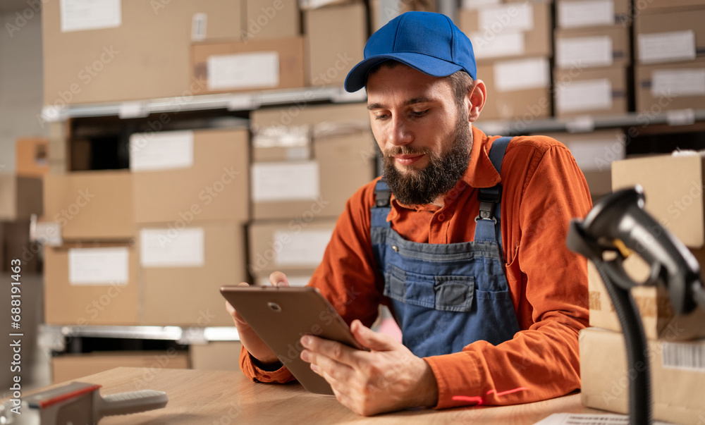 A warehouse worker conducting an inventory uses a digital tablet while sitting at a table in work clothes. - obrazy, fototapety, plakaty 