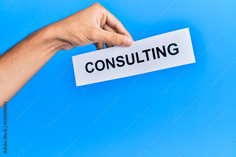 Hand of caucasian man holding paper with consulting word over isolated blue background Hand of caucasian man holding paper with consulting word over isolated blue background