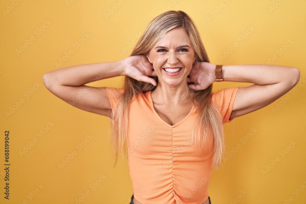 Young woman standing over yellow background relaxing and stretching, arms and hands behind head and neck smiling happy Young woman standing over yellow background relaxing and stretching, arms and hands behind head and neck smiling happy