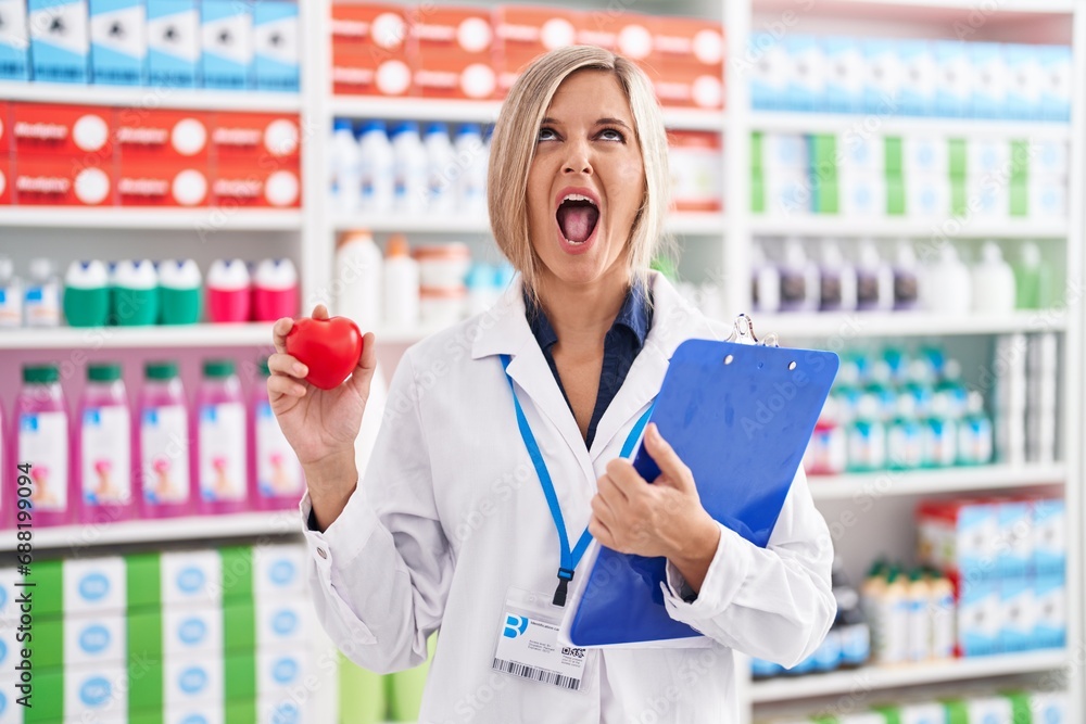 Young woman working at pharmacy drugstore holding heart angry and mad screaming frustrated and furious, shouting with anger looking up.